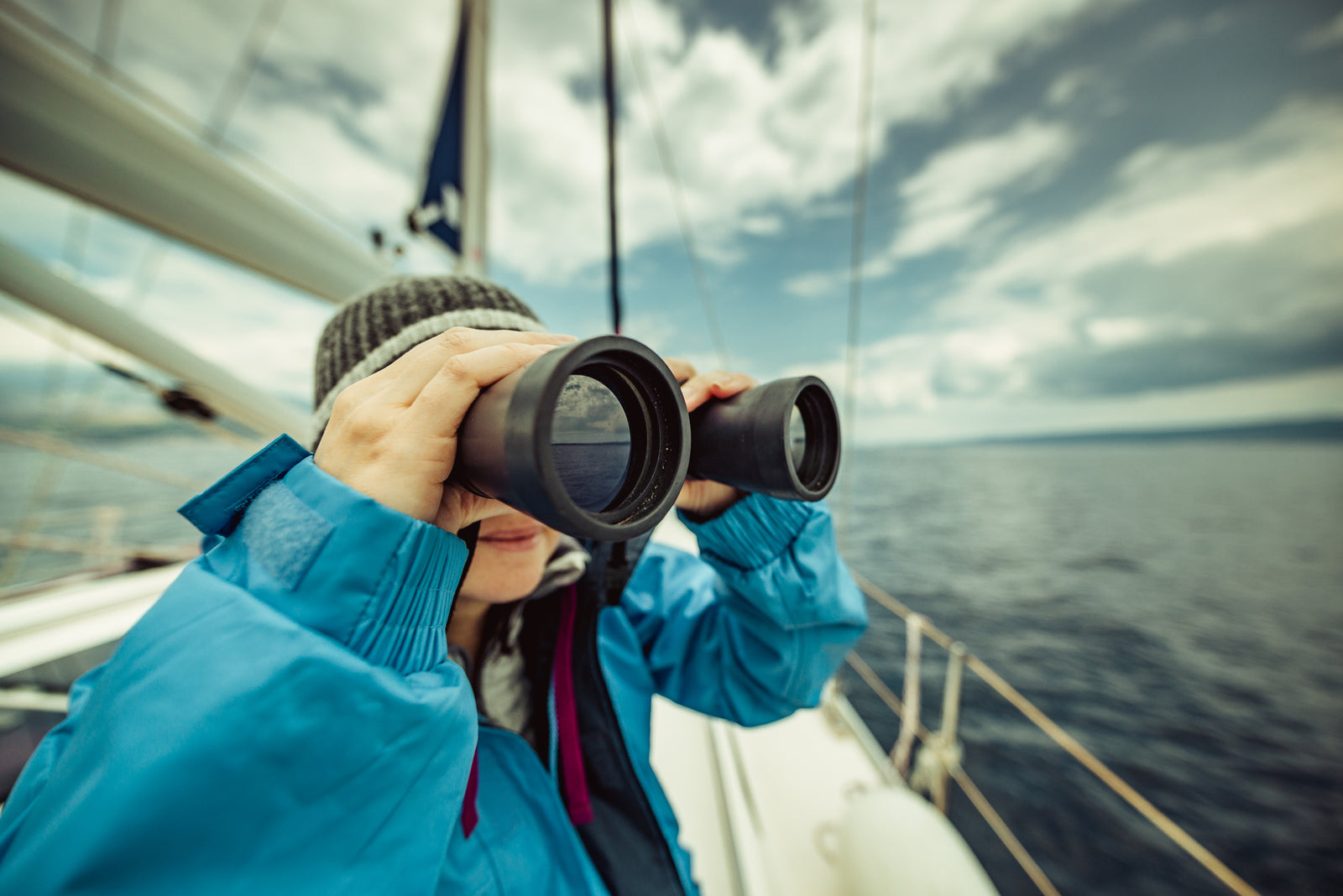 Woman in a waterproof jacket scanning the horizon with marine binoculars from a yacht deck