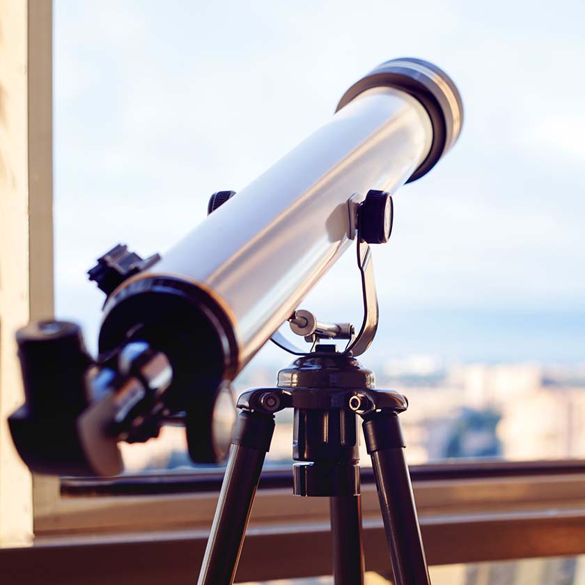 A telescope set up by a window with a cityscape in the background, ready for urban stargazing